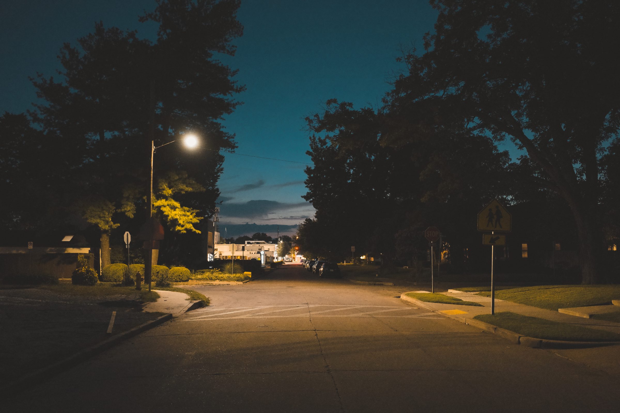 An empty residential intersection at dusk, one streetlight lit, downtown a few blocks ahead